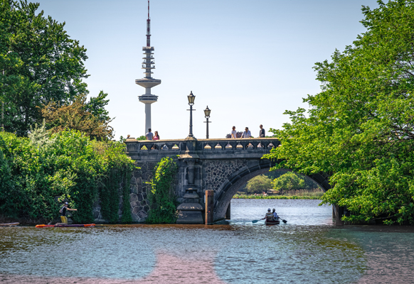 Personen fahren in einem Ruderboot auf der Alster unter einer Brücke hindurch in Hamburg.