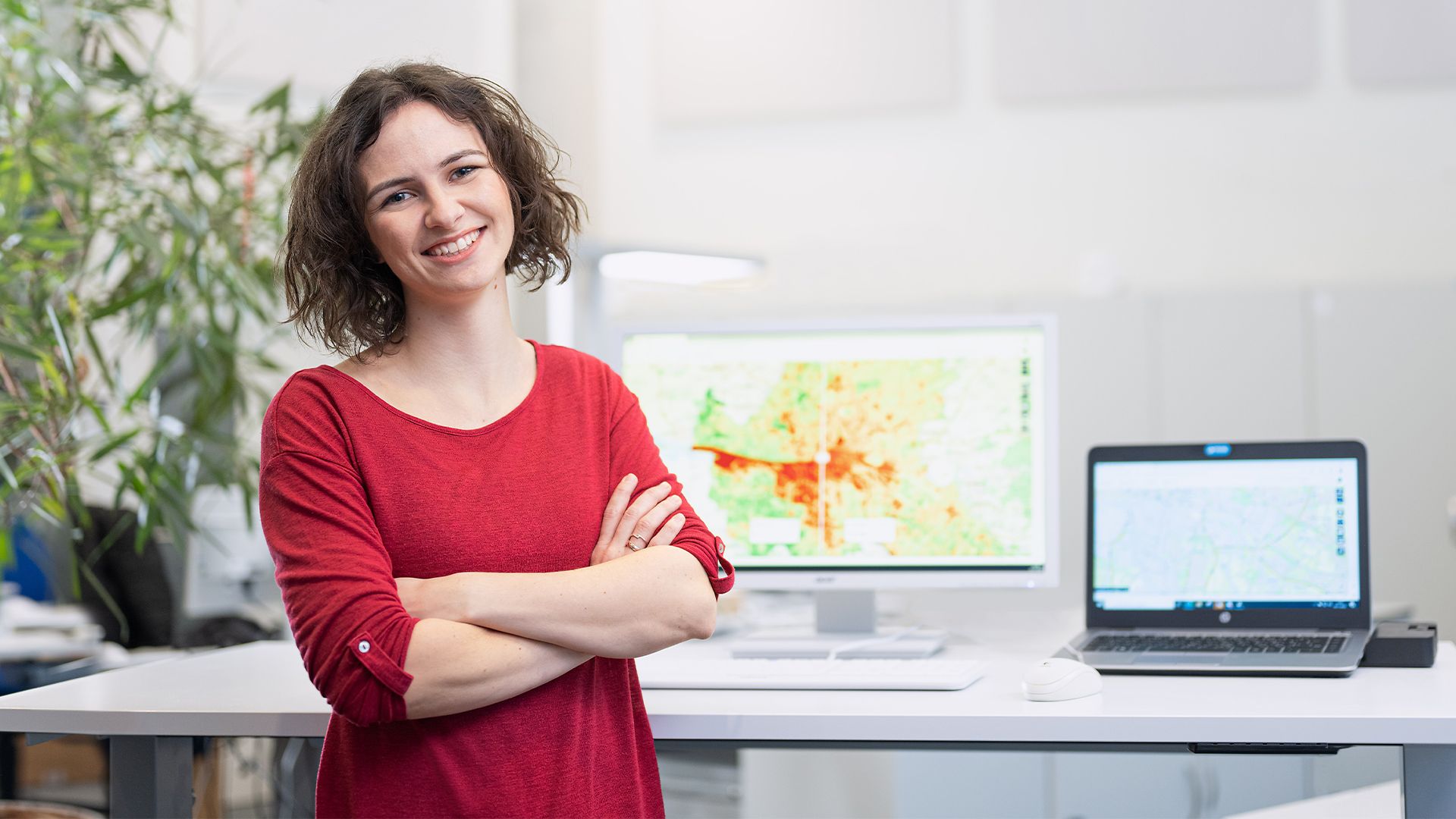 Eine Geoinformatikerin steht vor ihrem Schreibtisch, im Hintergrund sind digitale Karten von Hamburg zu sehen.